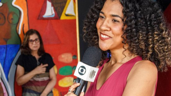 Jornalista Beatriz Oliveira, usa uma camisa rosa e segura um microfone durante reportagem para a Globo