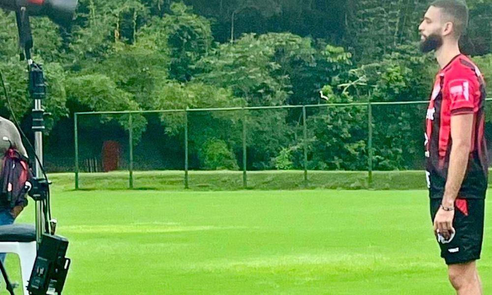 Imagem de um campo verde aberto ao fundo, equipamento de fotografia à direita e um homem branco vestindo uma camisa de time de futebol em vermelho e preto do lado direito