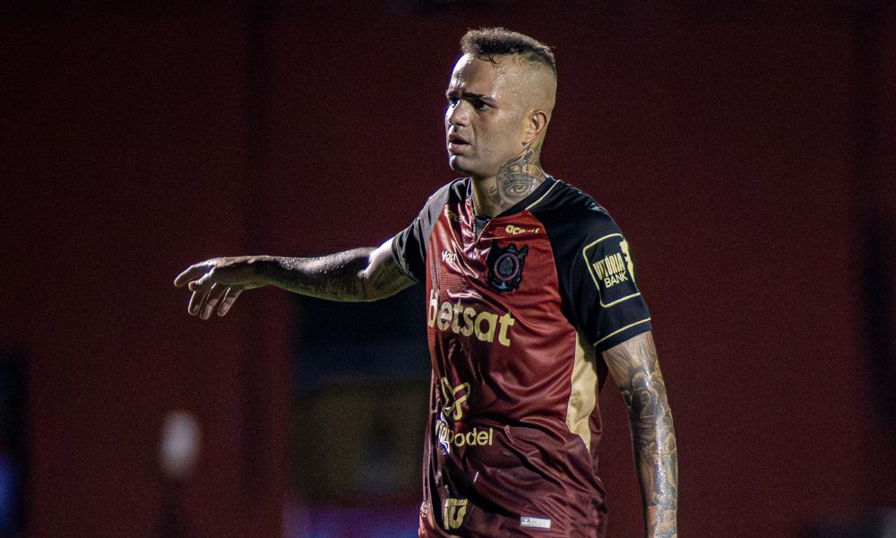 Homem branco com camisa vermelha com detalhes em preto e dourado, cenando para o lado