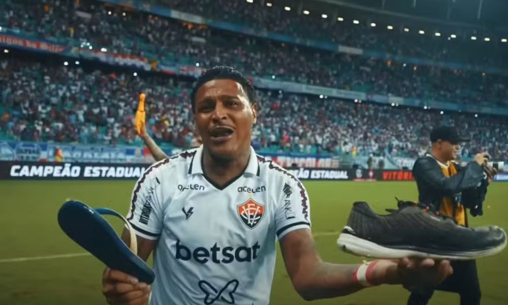 Foto do jogador Rodrigo Andrade com uniforme branco do Vitória segurando uma sandália azul e branca e um tênis preto