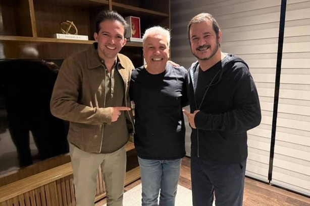 Três homens estão posando juntos em um ambiente interno. O homem à esquerda está usando uma jaqueta clara e calças escuras, apontando para o homem do meio, que está vestindo uma camiseta preta. O homem à direita está usando um conjunto de moletom escuro e também está apontando para o homem do meio. Ao fundo, há uma estante de madeira com alguns objetos decorativos e uma televisão. Rubinho Nunes, Sikêra Júnior e Rueda reunidos em São Paulo para discutir filiação partidária (Foto: Divulgação)